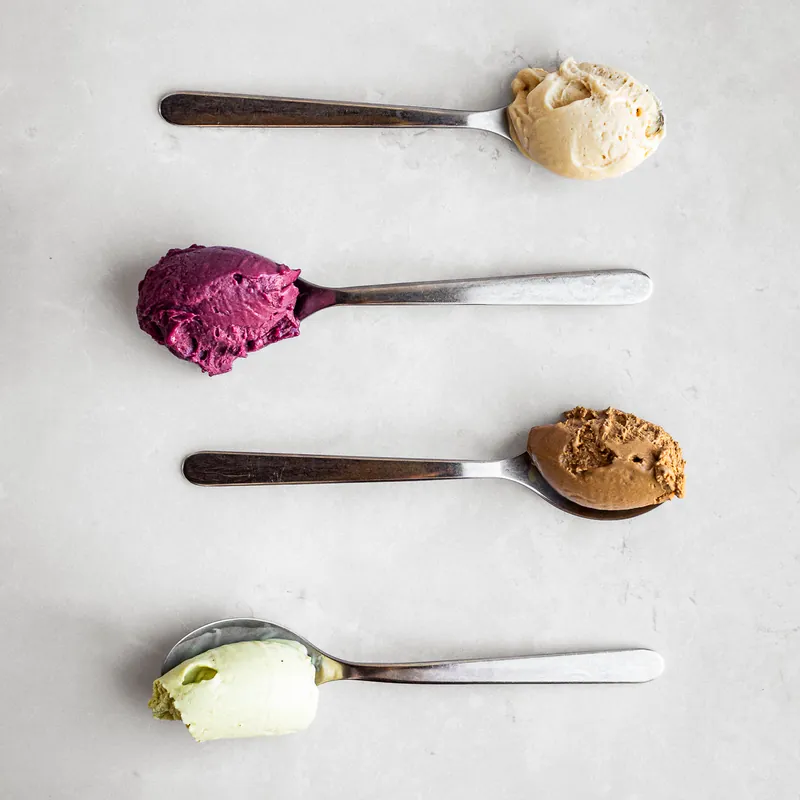 Quattro cucchiai di gelato artigianale allineati su un piano chiaro: crema, frutti di bosco, caffè e pistacchio.