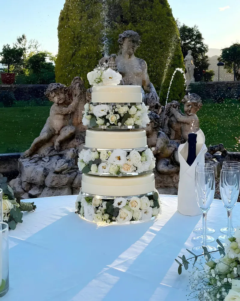 Torta nuziale a tre piani bianca decorata con rose bianche, allestita all'aperto davanti a una fontana con statue.
