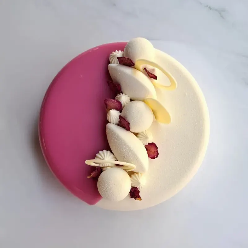 Vista dall'alto della torta La rosa: glassa metà rosa e metà bianca, con meringhe, sfere di cioccolato bianco e petali di rosa essiccati.