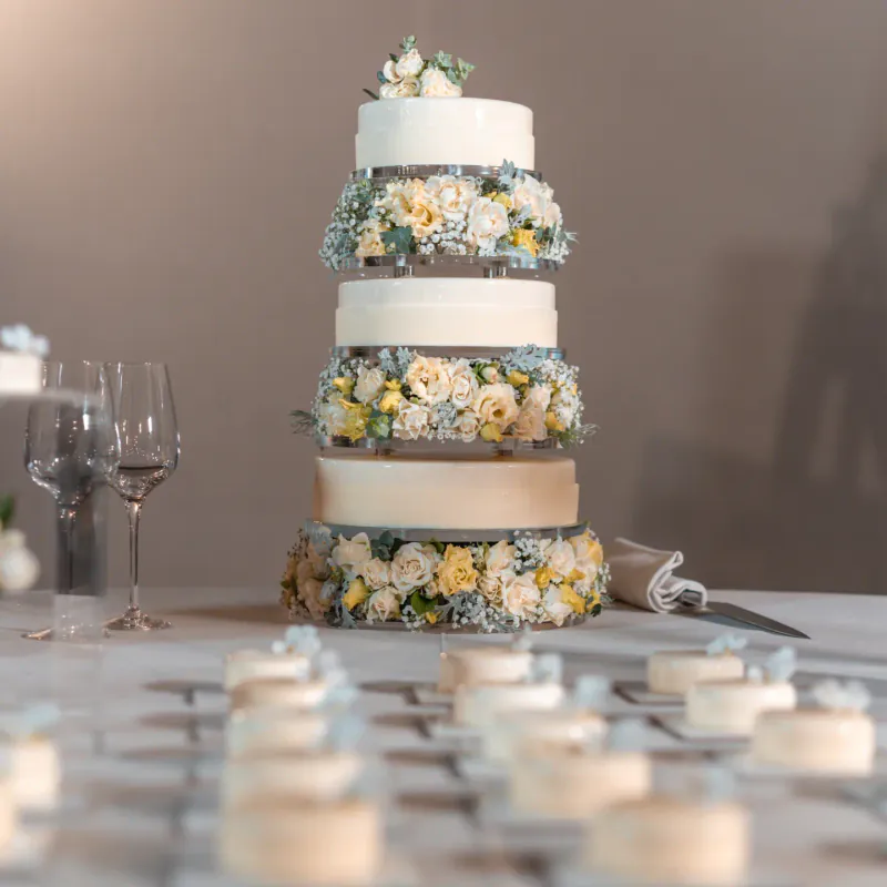Torta nuziale a tre piani bianca decorata con fiori chiari, con bicchieri da vino e mignon sfocati in primo piano.