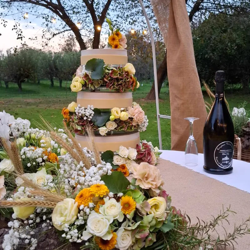 Torta nuziale a tre piani con ortensie, rose e gerbere arancioni, su un tavolo in iuta all'aperto fra gli alberi illuminati.