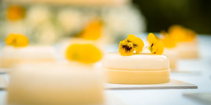 Monoporzioni di semifreddo decorate con viole del pensiero gialle, con la torta nuziale sfocata sullo sfondo.